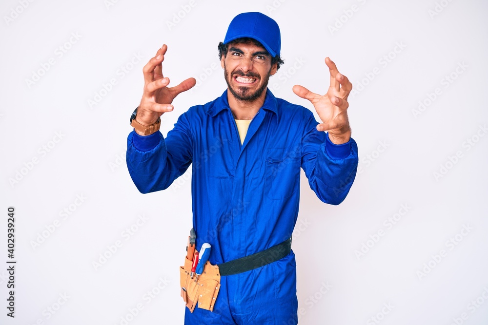 Handsome young man with curly hair and bear weaing handyman uniform shouting frustrated with rage, hands trying to strangle, yelling mad
