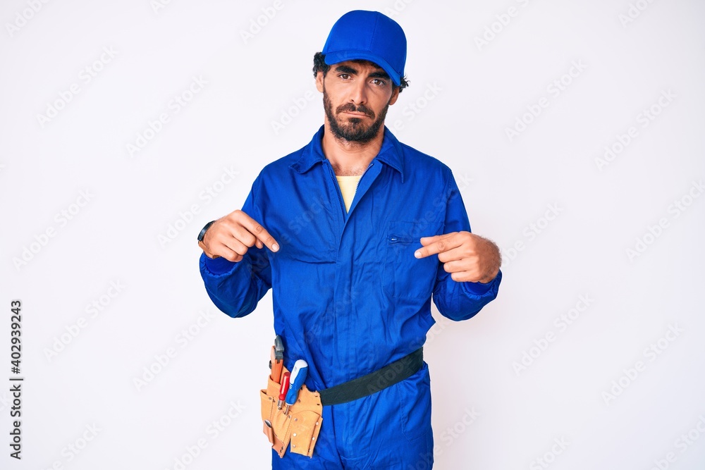 Handsome young man with curly hair and bear weaing handyman uniform pointing down looking sad and upset, indicating direction with fingers, unhappy and depressed.