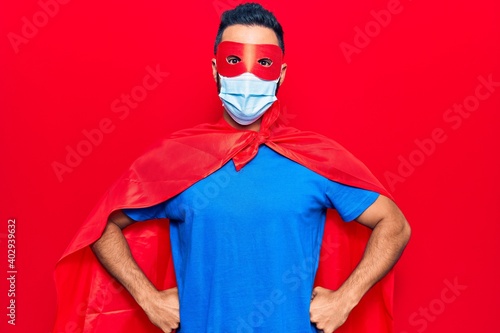 Young hispanic man wearing super hero costume and medical mask looking positive and happy standing and smiling with a confident smile showing teeth