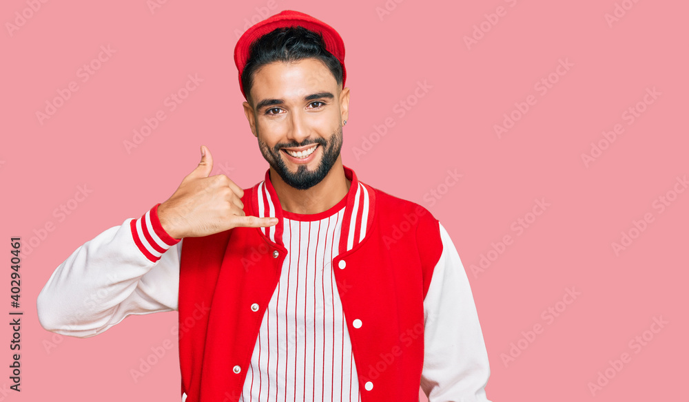 Young man with beard wearing baseball uniform smiling doing phone gesture with hand and fingers like talking on the telephone. communicating concepts. Young man with beard wearing baseball uniform smiling doing phone gesture with hand and fingers like talking on the telephone. communicating concepts.