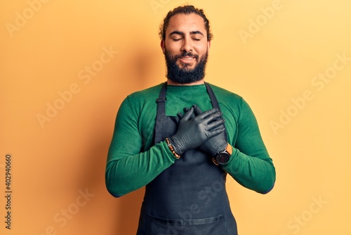 Young arab man wearing barber apron smiling with hands on chest with closed eyes and grateful gesture on face. health concept.