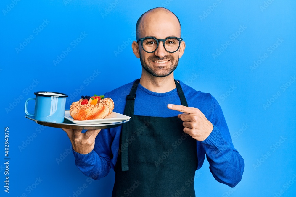 Young hispanic man wearing waiter apron holding tray with breakfast smiling happy pointing with hand and finger