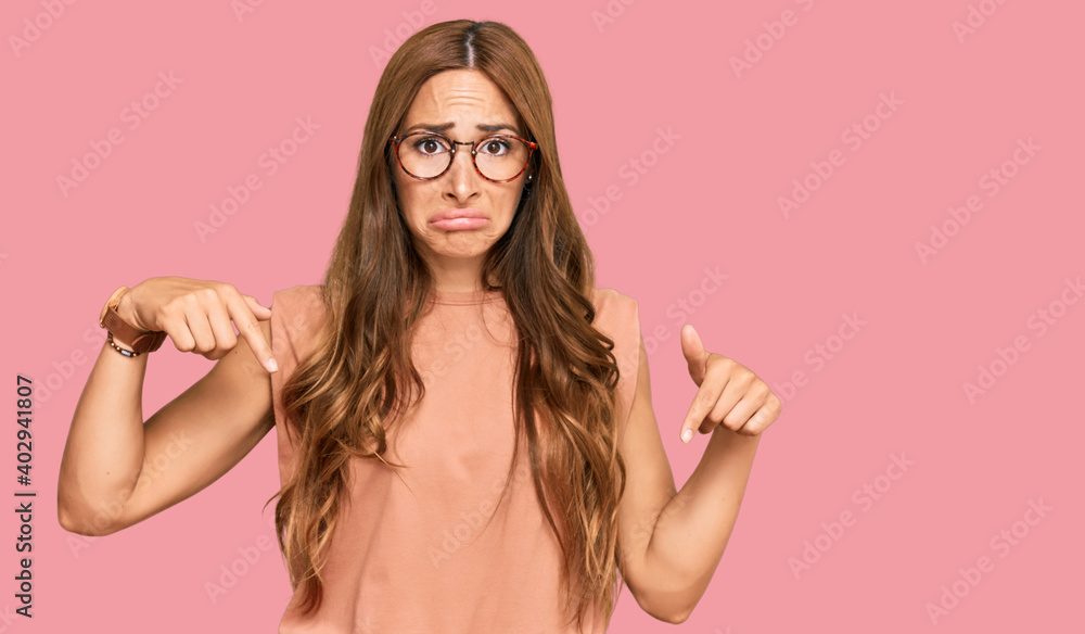 Young hispanic woman wearing casual clothes and glasses pointing down looking sad and upset, indicating direction with fingers, unhappy and depressed. Young hispanic woman wearing casual clothes and glasses pointing down looking sad and upset, indicating direction with fingers, unhappy and depressed.