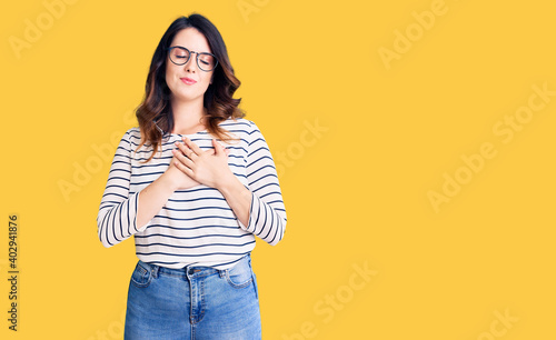 Beautiful young brunette woman wearing casual clothes and glasses smiling with hands on chest with closed eyes and grateful gesture on face. health concept.