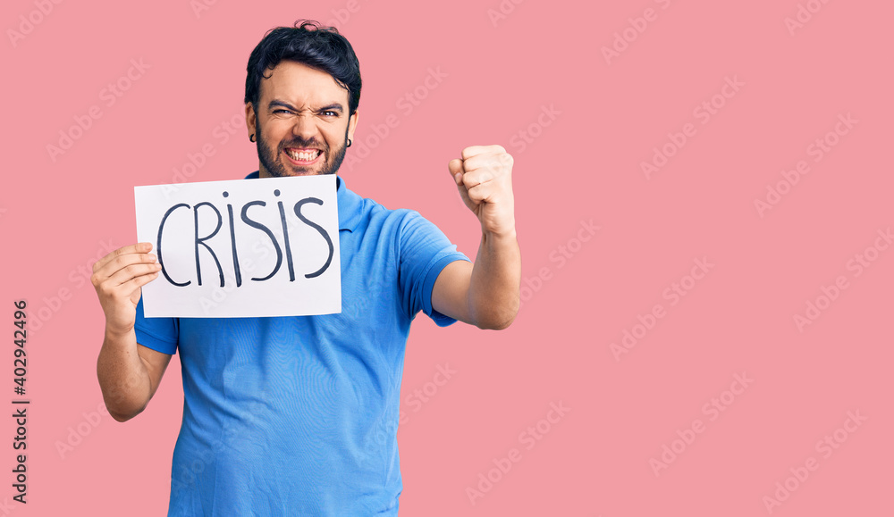 Young hispanic man holding crisis message paper annoyed and frustrated shouting with anger, yelling crazy with anger and hand raised Young hispanic man holding crisis message paper annoyed and frustrated shouting with anger, yelling crazy with anger and hand raised
