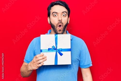 Young hispanic man holding gift scared and amazed with open mouth for surprise, disbelief face © Krakenimages.com