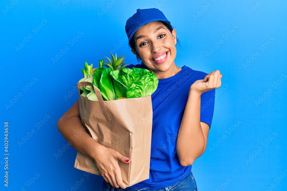 Beautiful brunette woman wearing courier uniform with groceries bag screaming proud, celebrating victory and success very excited with raised arm Beautiful brunette woman wearing courier uniform with groceries bag screaming proud, celebrating victory and success very excited with raised arm