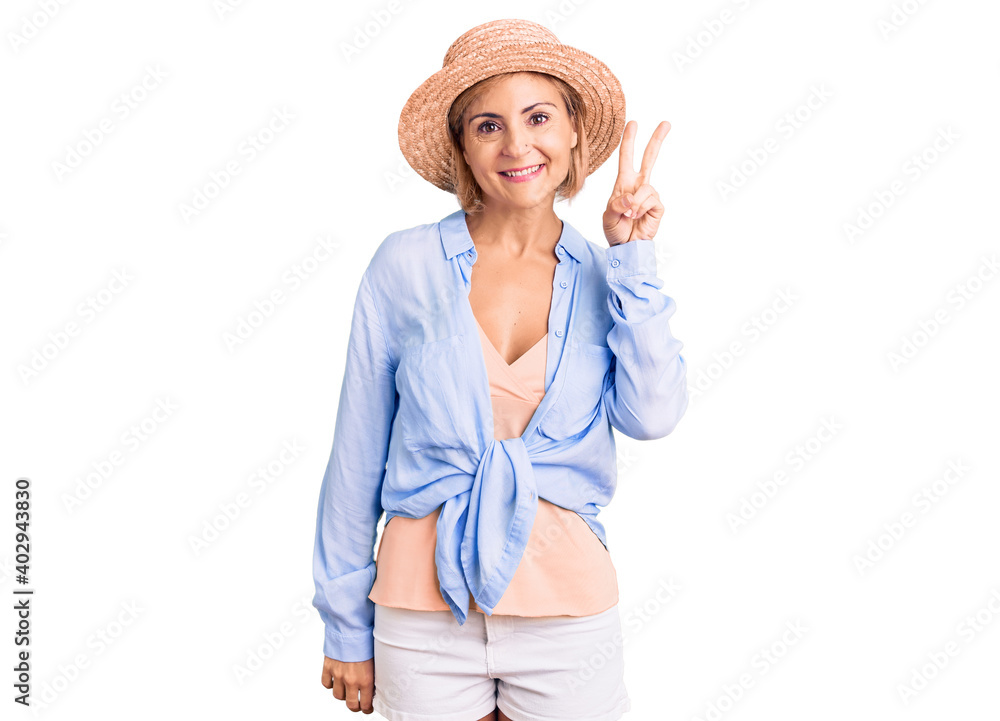 Young blonde woman wearing summer hat smiling with happy face winking at the camera doing victory sign. number two. Young blonde woman wearing summer hat smiling with happy face winking at the camera doing victory sign. number two.