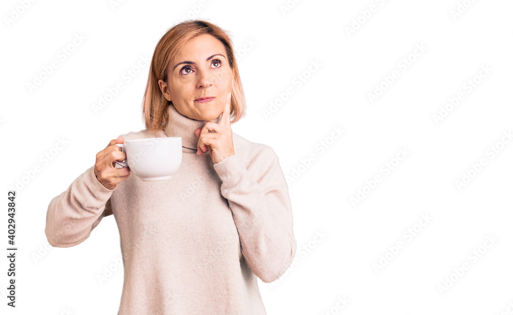 Young blonde woman holding coffee serious face thinking about question with hand on chin, thoughtful about confusing idea Young blonde woman holding coffee serious face thinking about question with hand on chin, thoughtful about confusing idea