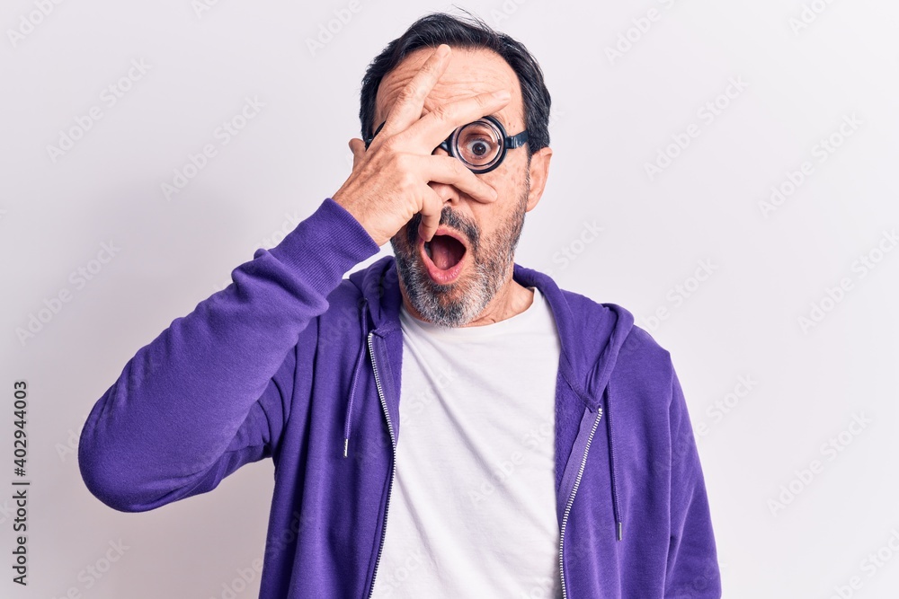 Middle age handsome freaky man wearing dumb glasses over isolated white background peeking in shock covering face and eyes with hand, looking through fingers afraid