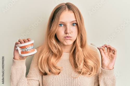 Beautiful young caucasian girl holding invisible aligner orthodontic and braces puffing cheeks with funny face. mouth inflated with air, catching air.