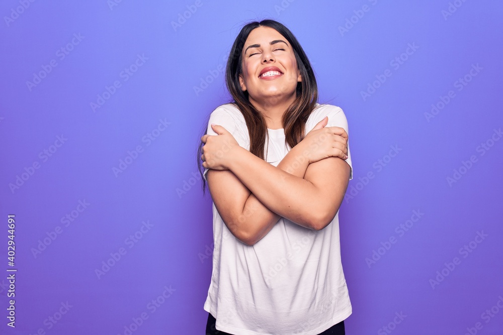 Young beautiful brunette woman wearing casual t-shirt standing over purple background hugging oneself happy and positive, smiling confident. Self love and self care Young beautiful brunette woman wearing casual t-shirt standing over purple background hugging oneself happy and positive, smiling confident. Self love and self care