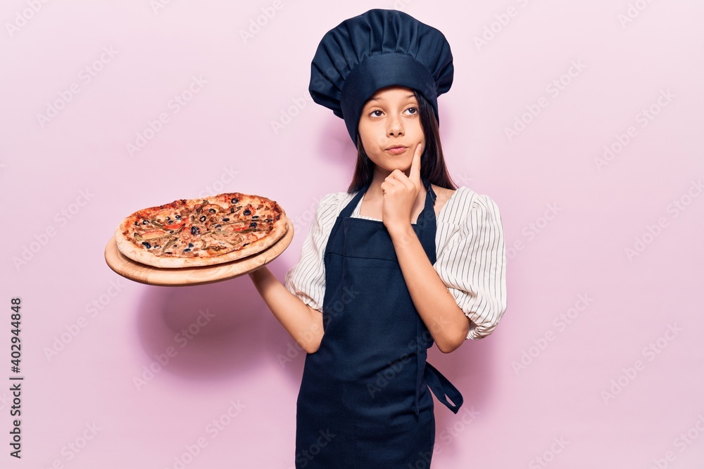 Beautiful child girl wearing cooker uniform holding italian pizza serious face thinking about question with hand on chin, thoughtful about confusing idea Beautiful child girl wearing cooker uniform holding italian pizza serious face thinking about question with hand on chin, thoughtful about confusing idea