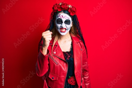 Woman wearing day of the dead costume over red angry and mad raising fist frustrated and furious while shouting with anger. rage and aggressive concept.