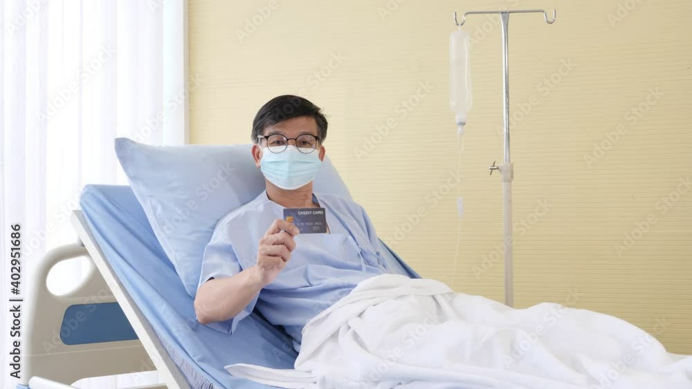 Asian male patient on hospital bed and wearing protective hygienic mask to prevent infection showing a credit card mockup for medical expenses. health insurance, Life Insurance and financial planning Asian male patient on hospital bed and wearing protective hygienic mask to prevent infection showing a credit card mockup for medical expenses. health insurance, Life Insurance and financial planning