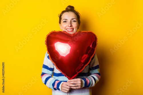 Young woman in a sweater holds an air balloon heart on a yellow background. Valentine's day composition © Alex