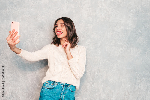 Young beautiful woman taking selfie photos. Trendy smiling model in casual summer jeans clothes. Positive female with red lips taking selfie photos. She posing near gray wall in studio. Shows tongue