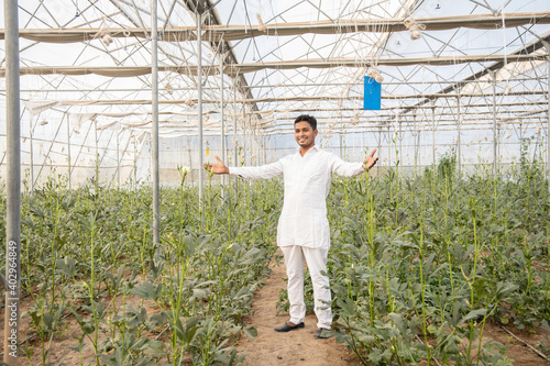 Young indian farmer standing with open arms at his poly house or greenhouse, agriculture business and rural prosperity concept. man wearing white kurta pajama cloths, copy space.