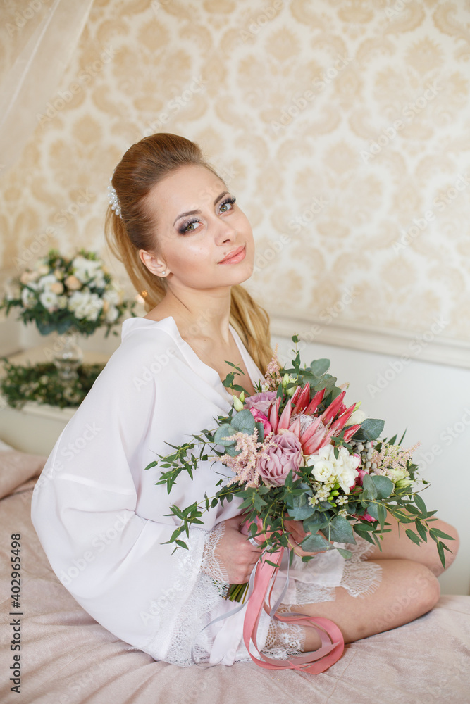 Pretty young Bride. Blonde-haired woman with wedding hair-style with a long tail. Boudoir morning of the bride. Looking on her bouquet Pretty young Bride. Blonde-haired woman with wedding hair-style with a long tail. Boudoir morning of the bride. Looking on her bouquet