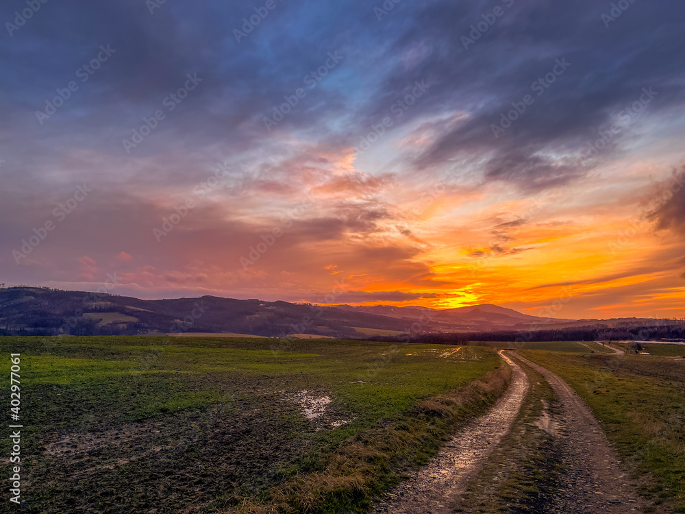 A dirt road leading around a field during an orange sunset on the horizon of the setting sun behind the hills.