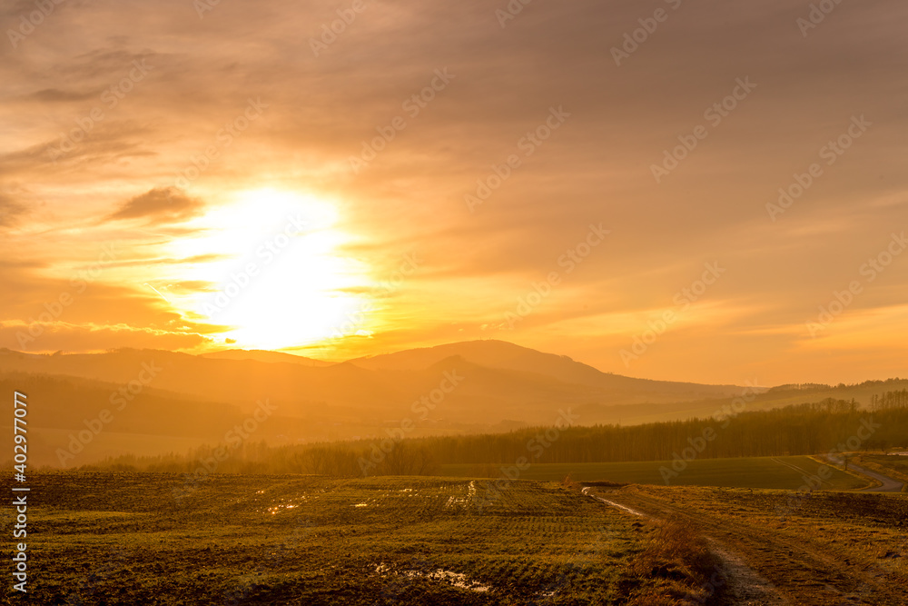 A dirt road leading around a field during an orange sunset on the horizon of the setting sun behind the hills. A dirt road leading around a field during an orange sunset on the horizon of the setting sun behind the hills.