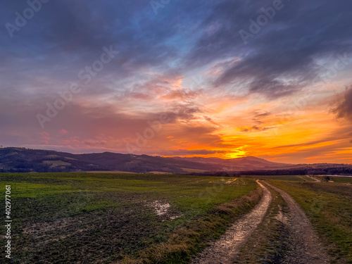 A dirt road leading around a field during an orange sunset on the horizon of the setting sun behind the hills.