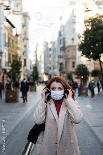 Beautiful girl in fashionable clothes holds and wears disposable mask at street. New normal lifestyle concept.