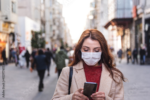 Beautiful girl wearing protective medical mask and fashionable clothes uses smart phone at street. New normal lifestyle concept.