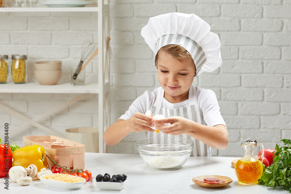 little boy in chef hat and an apron cooking dough for pizza in the kitchen. child cooking with eggs little boy in chef hat and an apron cooking dough for pizza in the kitchen. child cooking with eggs