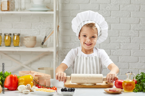 little child boy in chef hat and an apron roll a dough for pizza in the kitchen