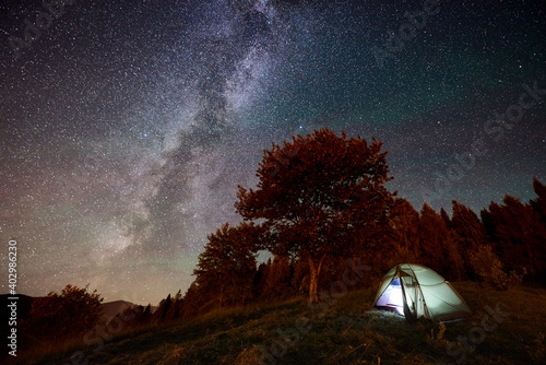 Tourist camping near forest at night. Illuminated tent under amazing night sky full of stars and Milky way. On the background beautiful starry sky and mountains
