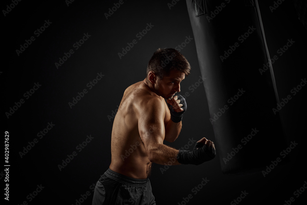 Aggressive shirtless boxer punching in boxing bag on black background