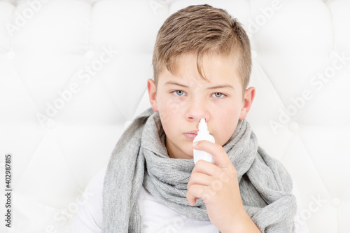 Sick teen boy spraing his nose with medicine while sitting on bed at home. An unhealthy child at home. Cold season. photo