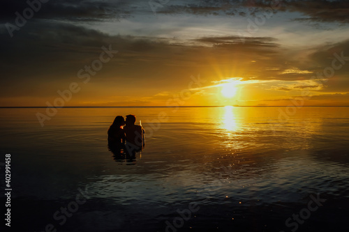 Young couple kissing and having a beer in the water during a very colorful sunset on the beach