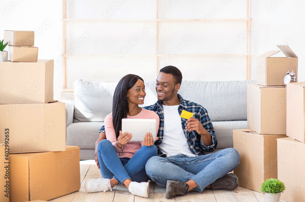 Black couple shopping for household goods online on moving day, sitting with laptop and credit card among carton boxes Black couple shopping for household goods online on moving day, sitting with laptop and credit card among carton boxes