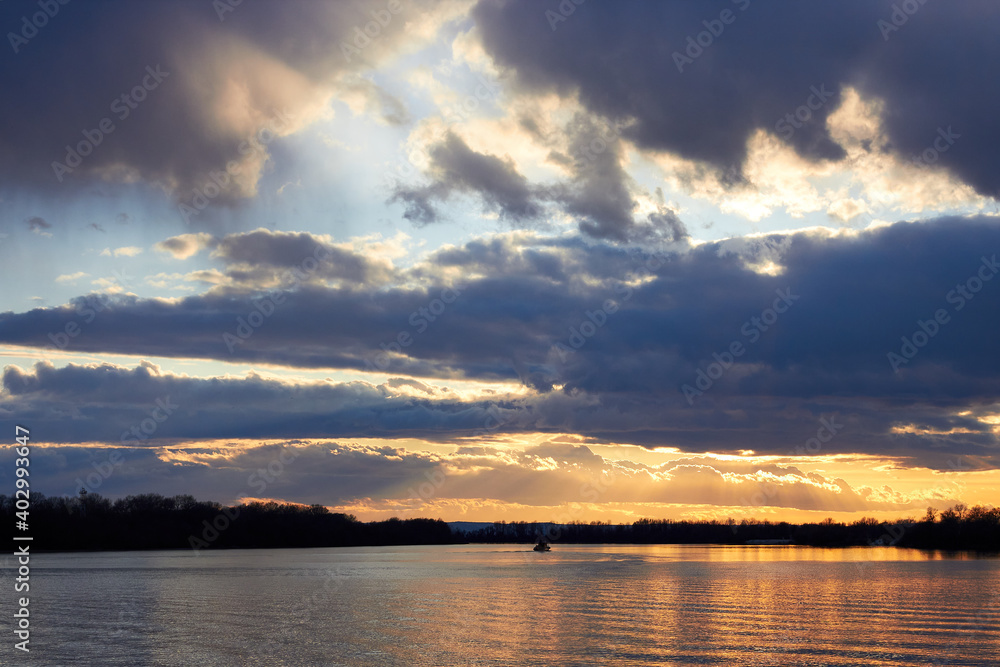 Landscape view of the river in the evening time, with wonderful blue cloudy sky during the yellow sunset lighting and a barge (ship) on the horizon Landscape view of the river in the evening time, with wonderful blue cloudy sky during the yellow sunset lighting and a barge (ship) on the horizon