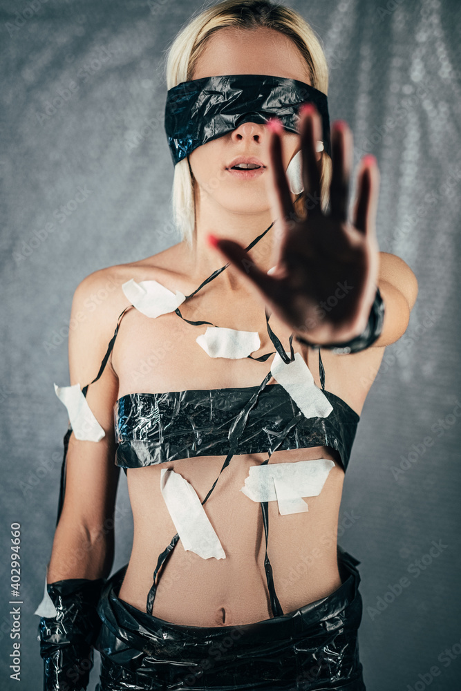 Stop abusing. Conceptual female portrait. Mental power. Domestic violence. Human rights. Woman with taped eyes body showing hand up isolated on gray wrinkled plastic film.