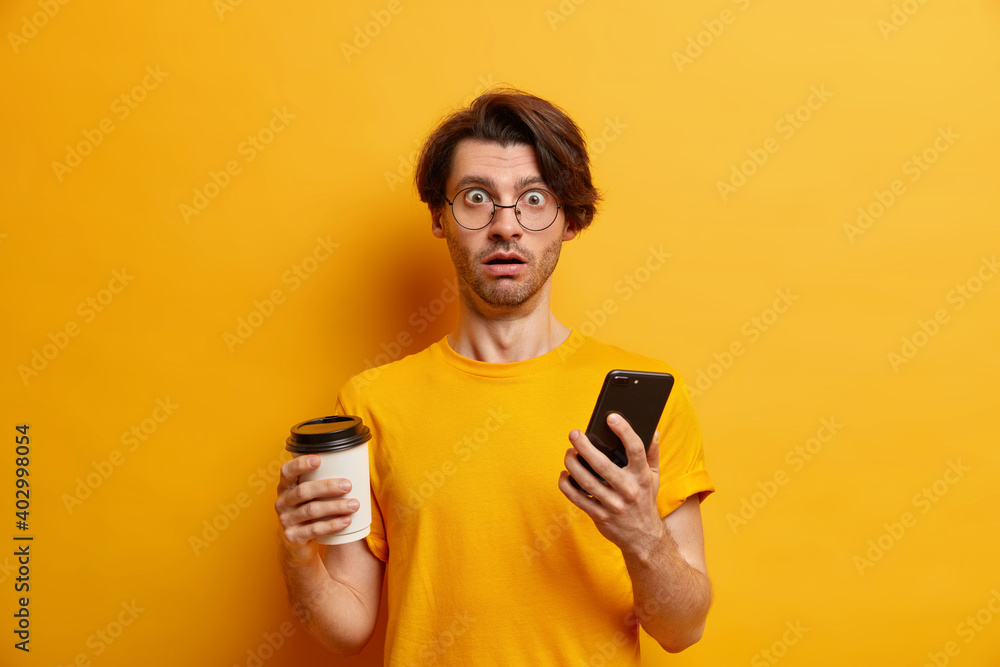 Shocked handsome guy reads unexpected message content stares stunned at camera holds disposable cup of coffee modern cellular wears round spectacles and t sirt isolated over yellow studio wall Shocked handsome guy reads unexpected message content stares stunned at camera holds disposable cup of coffee modern cellular wears round spectacles and t sirt isolated over yellow studio wall