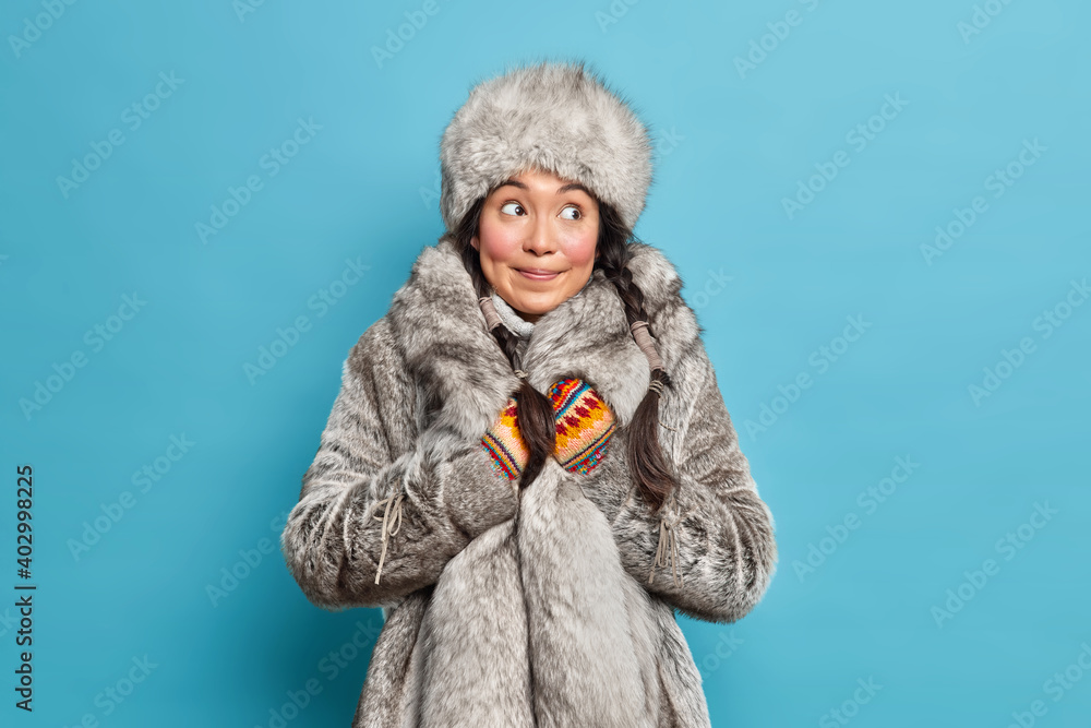 Thoughtful Inuit woman dressed in grey fur hat and coat knitted mittens wears warm winter clothes isolated over blue studio background. Scandinavian female wears outerwear lives in far north Thoughtful Inuit woman dressed in grey fur hat and coat knitted mittens wears warm winter clothes isolated over blue studio background. Scandinavian female wears outerwear lives in far north