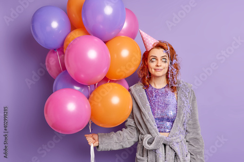 Funny birthday girl feels tired after noisy party smiles pleasantly concentrated above smeared with cream has festive mood holds multicolored balloons isolated over purple background. Holidays concept