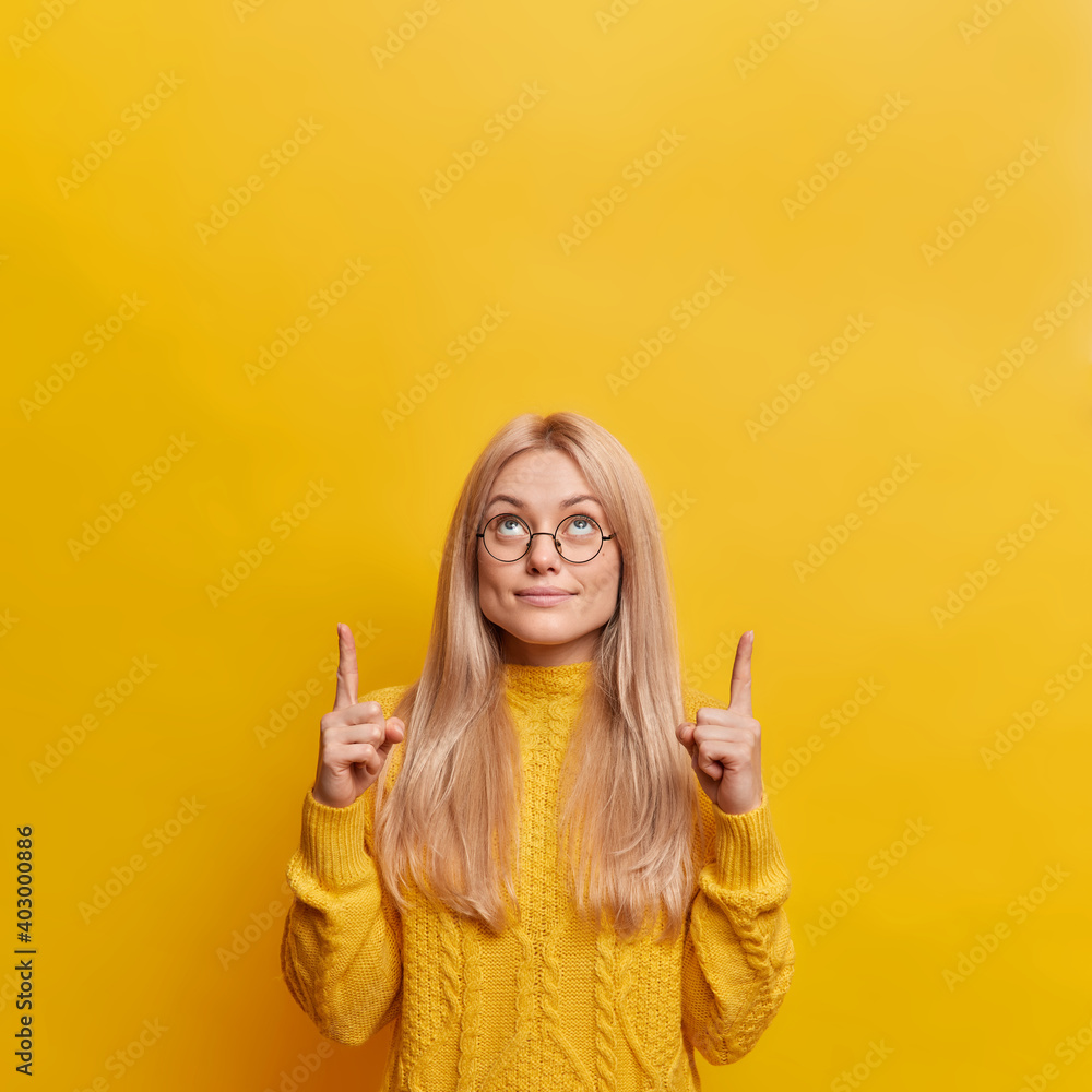 Good looking blonde woman indicates both index fingers upwards shows amazing offer dressed in casual sweater isolated over yellow background. Look at this promotion. Use copy space for your advert Good looking blonde woman indicates both index fingers upwards shows amazing offer dressed in casual sweater isolated over yellow background. Look at this promotion. Use copy space for your advert