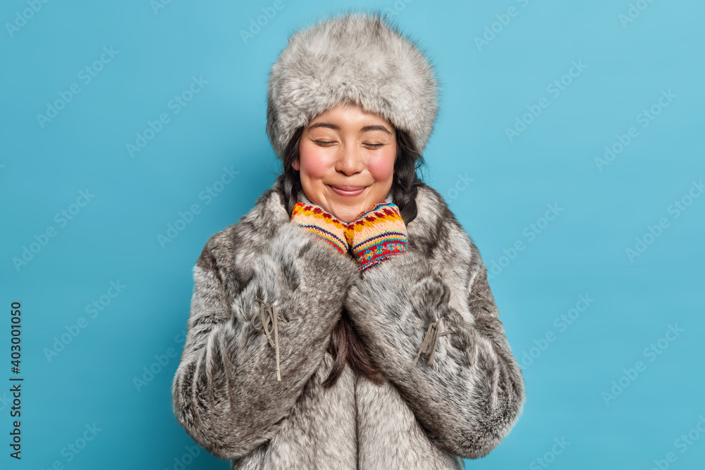 Dreamy satisfied young Asian woman closes eyes and smiles gently wears grey fur hat and coat has romantic mood poses against blue background. Eskimo female from north. Arctic aboriginal indoor Dreamy satisfied young Asian woman closes eyes and smiles gently wears grey fur hat and coat has romantic mood poses against blue background. Eskimo female from north. Arctic aboriginal indoor