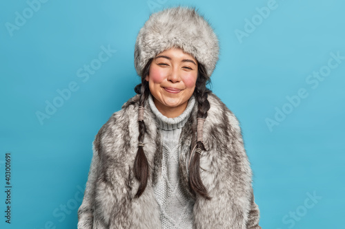 Winter clothes. Pleased Inuit woman with two pigtails smiles gently wears fur hat knitted warm sweater and coat poses against blue background. Happy nenets girl indoor. Aboriginal from north photo