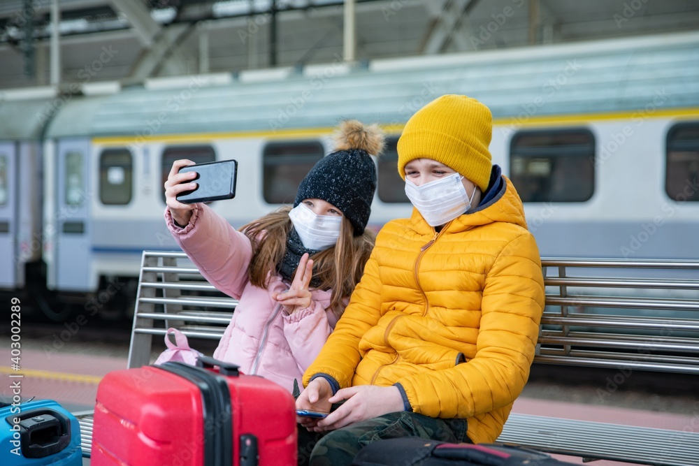 Two children wearing protective masks are sitting on the platform waiting for the train. The girl takes a selfie. Two children wearing protective masks are sitting on the platform waiting for the train. The girl takes a selfie.