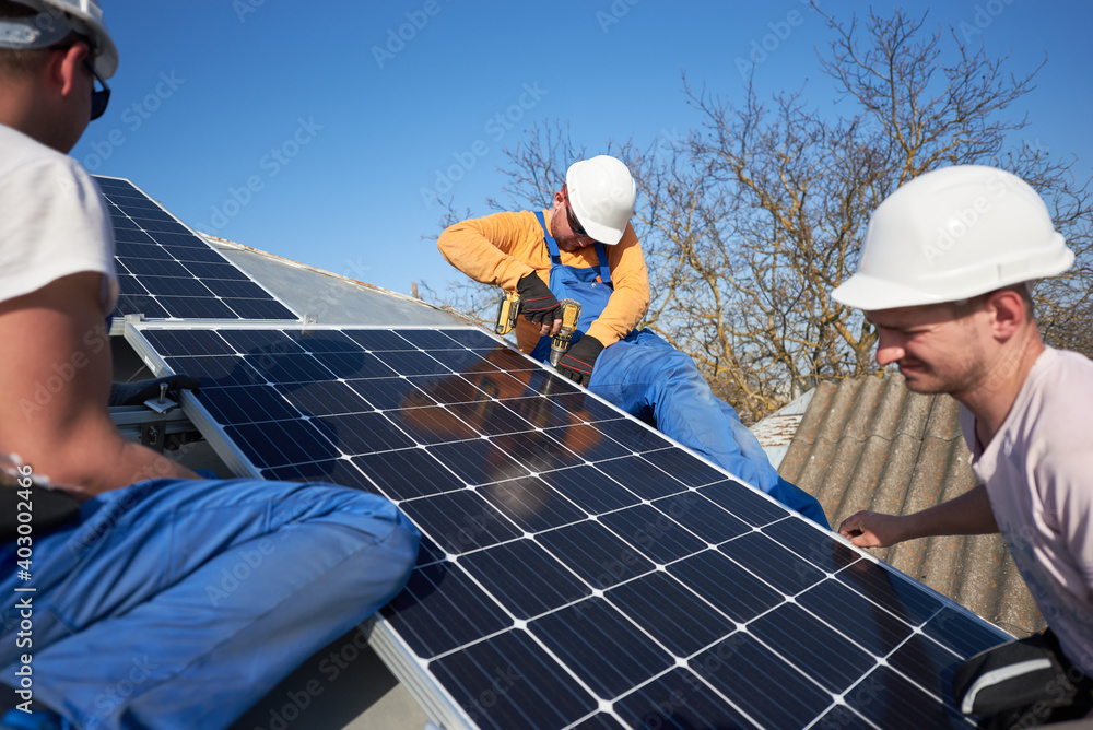 Male engineers installing solar photovoltaic panel system using screwdriver. Electricians mounting blue solar module on roof of modern house. Alternative energy ecological concept. Male engineers installing solar photovoltaic panel system using screwdriver. Electricians mounting blue solar module on roof of modern house. Alternative energy ecological concept.