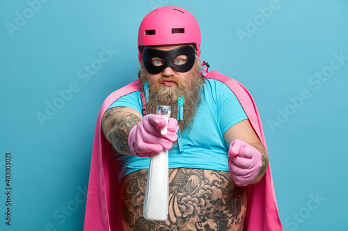 Strict serious bearded man clenches fist from ager sprays detergent at you wears pink helmet eye mask and cape pretends to be cleaning superhero washes everything in room poses against blue wall
