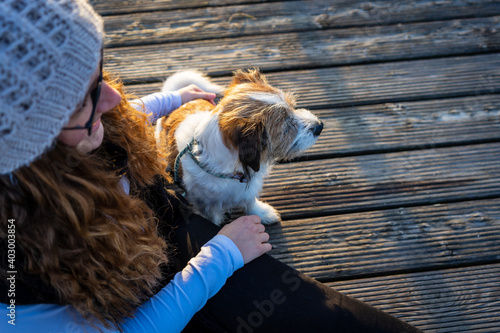 Junge Frau mit Wollmütze und langen roten Haaren und ein kleiner Terrier Hund sitzen auf einem Holzsteg. Draußen, Winter, Lifestyle.