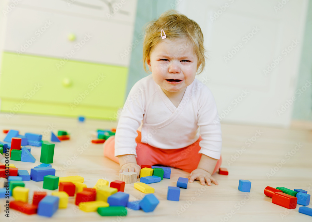 Upset crying baby girl with educational toys. Sad tired or hungry alone healthy child sitting near colorful different wooden blocks at home or nursery. Baby missing mother in daycare Upset crying baby girl with educational toys. Sad tired or hungry alone healthy child sitting near colorful different wooden blocks at home or nursery. Baby missing mother in daycare