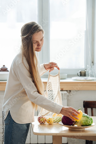 Gentle blonde woman in a sunlit room accurately placing vegetables on a dish and out of a net bag. At home in the kitchen.