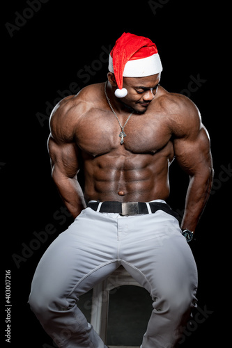 Black fitness model man sits on high bar chair on black background. Naked torso of very muscular man.